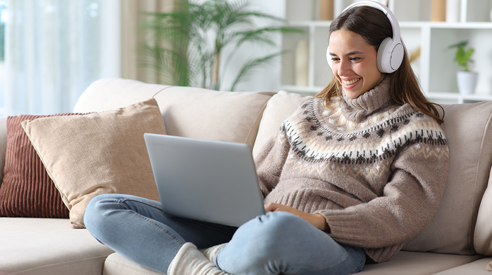 Happy woman in winter using laptop and headphone sitting on a couch at home